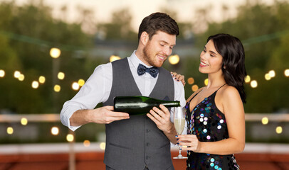 celebration and holidays concept - happy couple with bottle of non alcoholic champagne and wine glass at rooftop party over lights on background