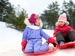 childhood, leisure and season concept - happy little girls on sleds outdoors in winter over snowy forest or park background