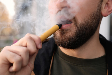 Handsome young man using disposable electronic cigarette outdoors, closeup