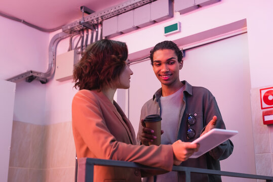 smiling african american technician with paper cup pointing at digital tablet while working with colleague in data center