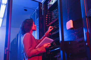 young technician holding digital tablet while checking wires in server in data center
