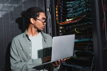 african american engineer holding laptop while looking at wires in server