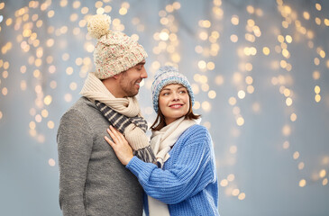 people and winter holidays concept - happy couple in knitted hats and scarves over christmas lights background