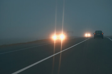 Headlights of oncoming cars driving in fog. Driving in fog, car headlights at foggy evening. - obrazy, fototapety, plakaty