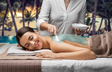 Obraz na płótnie Canvas wellness, beauty and cosmetology concept - beautiful young woman lying with closed eyes and cosmetologist applying clay mask by spatula at spa over tropical beach background in french polynesia