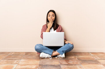 Young mixed race woman with a laptop sitting on the floor covering mouth with hand