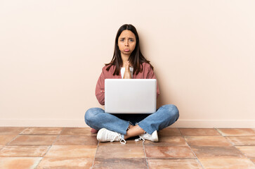 Young mixed race woman with a laptop sitting on the floor keeps palm together. Person asks for something