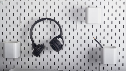 Black wireless headphones hang on a hook on a white wall near a desktop with shelves for stationery...