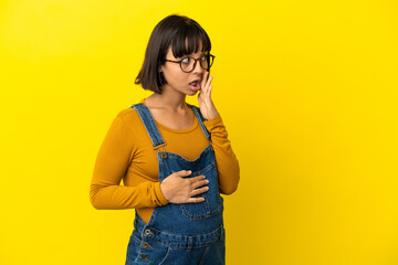 Young pregnant woman over isolated yellow background whispering something with surprise gesture while looking to the side