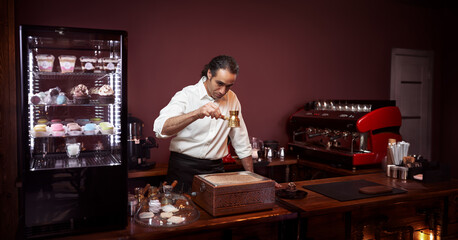 Young cheerful barista wearing black apron and white shirt while preparing turkish coffee in a modern coffee shop