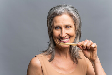 Headshot of smiling senior gray-haired woman with bamboo toothbrush brushing her white teeth isolated on gray background, everyday care routine. Dental care and oral hygiene