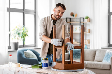 furniture renovation, diy and home improvement concept - happy smiling man sanding old wooden table or chair with sponge