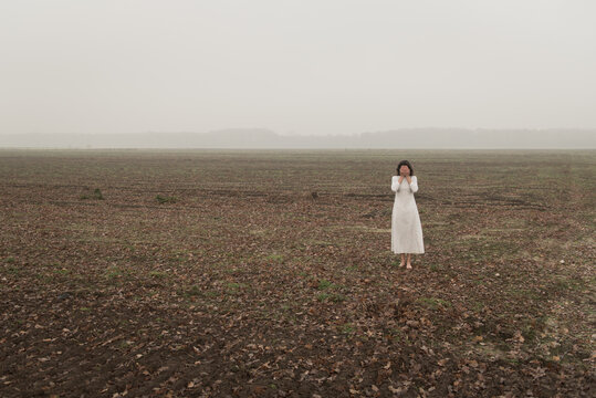 Fine art portrait of woman in white dress standing alone in bare field on misty cold day
