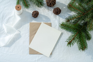 Blank greeting card, invitation card mockup.Christmas decoration scene in sunlight.Paper,  envelope, ribbon, fir branches, wooden balls.White linen background.Top view, flat lay.Winter wedding concept