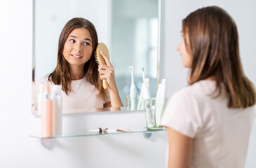 beauty and people concept - teenage girl looking to mirror and brushing wet hair with comb at home...