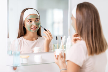 Obraz na płótnie Canvas beauty, skin care and people concept - teenage girl with brush applying clay mask to face and looking in mirror at bathroom