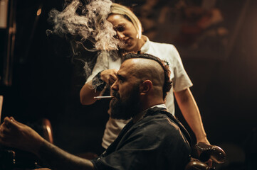 Young bearded man with a mohawk getting haircut by hairdresser and smoking a cigarette