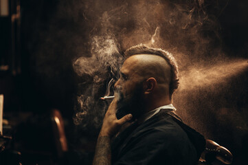 Young bearded man with a mohawk sitting at barbershop and smoking a cigarette