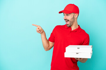 Delivery caucasian man picking up pizza boxes isolated on blue background pointing finger to the side and presenting a product