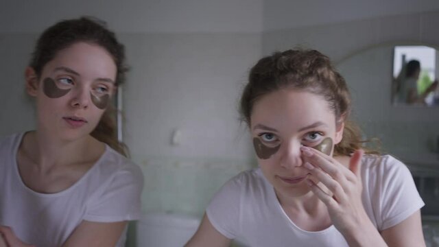 Front View Portrait Twin Sisters Fighting For Mirror Applying Moisturizing Eye Mask In Bathroom At Home. Slim Beautiful Caucasian Young Women Taking Care Of Facial Skin In The Morning. Beauty Routine