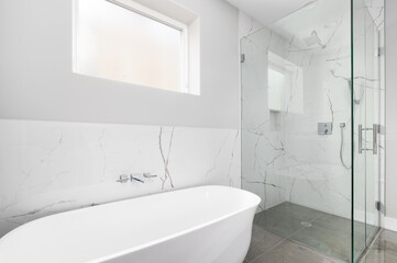 Fototapeta na wymiar A luxurious shower with glass and marble tiled walls. The shower has a built in shelf and chrome shower head. A white freestanding tub is below a frosted window.