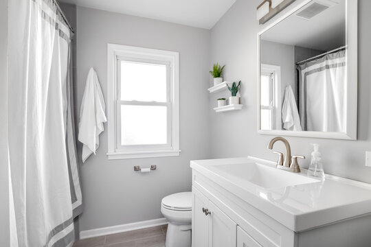 A Cozy White Bathroom With A Bronze Faucet On Marble Countertop, Plants On Floating Shelves, And Tiled Floor.