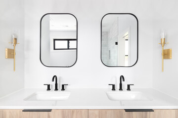 A beautifully renovated bathroom with a wood cabinet, black framed mirror and faucets, and gold lights mounted on the wall.