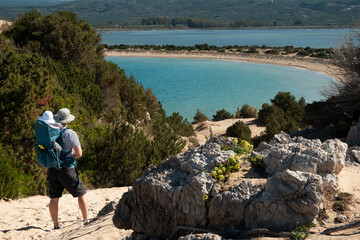 Man wearing sunhat carrying baby in backpack, hiking, travelling in beautiful place, on background of blue lagoon, sea, Voidokilia beach in Greece,Peloponnese.Trip with kid, beauty of nature concept