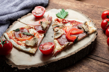 Pizza with ham and bacon, mushrooms and tomatoes on a slice of wood on a brown wooden background.