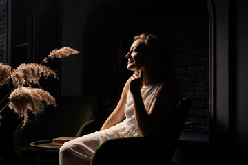 A young woman in a white dress sits thoughtfully in an armchair in a dark room. Solitude,...