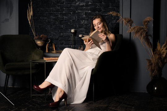 A young woman in a white dress is reading a book in a dark room.