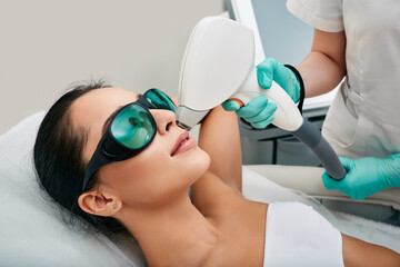 Attractive woman wearing protective glasses while laser hair removal and laser epilation to lips area on her face at a beauty salon - obrazy, fototapety, plakaty