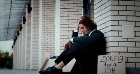 Depressed young man entrepreneur sitting on ground next to wall, looking anxious, frustrated, suffering from severe headache, thinking of financial problems, money loss.