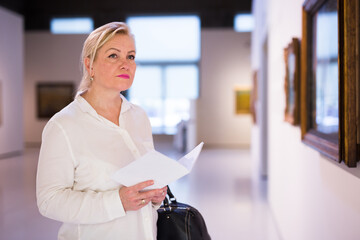 Senior female visitor holding guidebook standing at painting exhibition in art museum