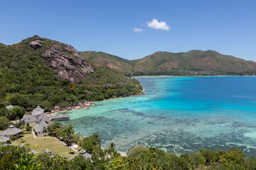 Panoramic view to Anse Petite Cour on Praslin island in the Seychelles from a high viewpoint. Bungalow hotel on a beach with a crystal blue lagoon, endless ocean on a summer day. Tropical landscape.
