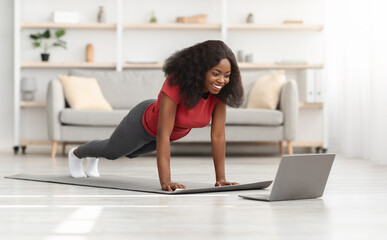 Fototapeta premium Positive african american athletic woman exercising at home, using laptop