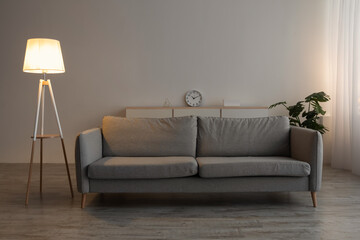 Sofa, clock, potted plant and glowing lamp in evening on gray wall background in office or living room