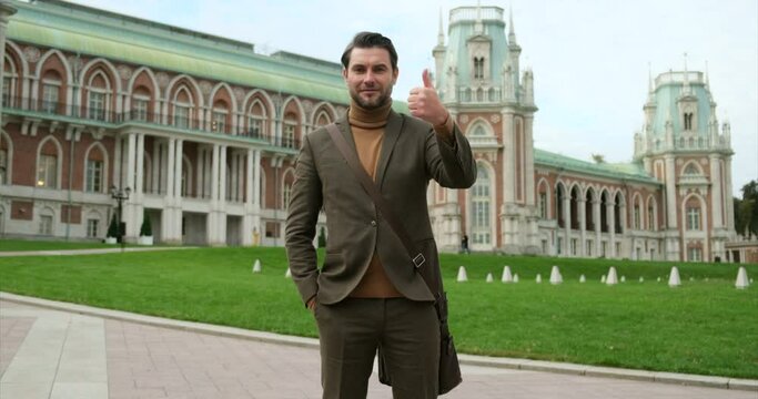 law graduate stands on the campus of a business school. businessman in an expensive stylish brown suit against the background of parliament show thumbs up. successful man in suit looking at the camera