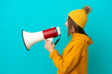 Young English woman wearing winter jacket isolated on blue background shouting through a megaphone