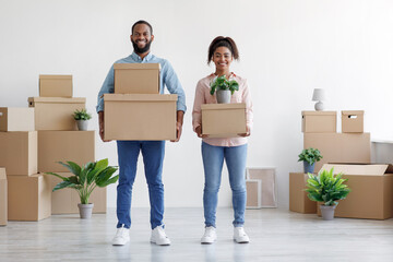 Fototapeta na wymiar Smiling young black couple carry cardboard boxes, potted plants in room with white walls interior