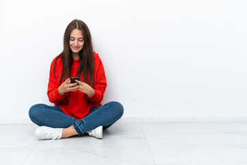 Young Ukrainian woman sitting on the floor isolated on white background sending a message with the mobile