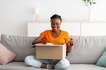 Full length of positive black lady opening cardboard package, receiving online store delivery at...