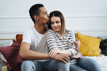 Obraz premium Portrait of happy young African Caucasian couple hug sitting on couch at home, smiling mixed race husband and wife spend time together, embrace relaxing on sofa. Healthy relationships concept