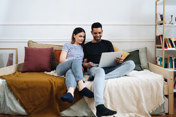 Fototapeta na wymiar Happy young mixed race couple enjoying time at home using computer sitting on sofa, black man and white woman having fun online shopping or surfing web relaxing on couch