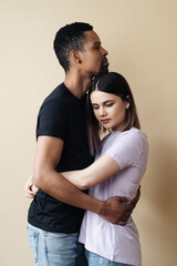 Portrait of happy multiracial couple hugging and posing together over beige background in studio, romantic guy cuddling his afro girlfriend