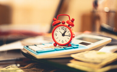 Um relógio despertador vermelho sobre uma calculadora azul com algumas cédulas de dólares americanos e um laptop na composição. Conceitos de finanças, economia, financiamento e tempo é dinheiro. - obrazy, fototapety, plakaty