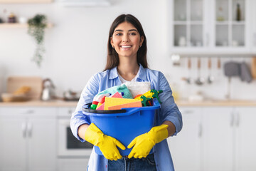 Young housewife holding bucket with cleaning supplies tools in kitchen