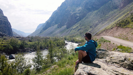 A man with glasses sits on the edge of a cliff wearing a blue anorak. Katu-Yaryk canyon Chulyshman Valley. Altai