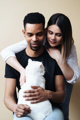 Lovely mixed-races young happy joyful couple male and female smiling in good mood resting with pet animal holding cat in hands, pet lover concept on beige background