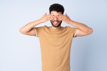 Young Moroccan man isolated on blue background covering eyes by hands and smiling